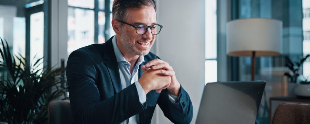 Homme souriant en costume, portant des lunettes, en visioconférence depuis un bureau lumineux.