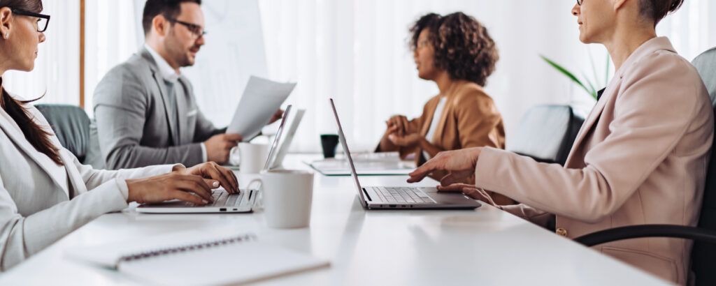Groupe de personnes en tenue professionnelle, travaillant sur des ordinateurs portables et échangeant autour d’une table de réunion dans un bureau lumineux.